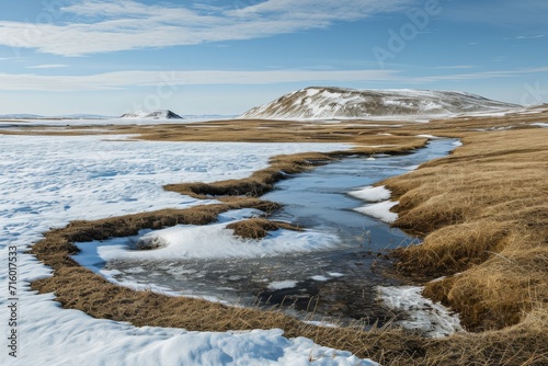 A tranquil stream winds its way through a frozen tundra, surrounded by towering mountains and icy rocks, under a winter sky filled with billowing clouds