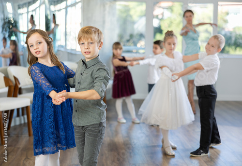 Happy little children in elegant dresses practicing waltz dance with teacher in school hall