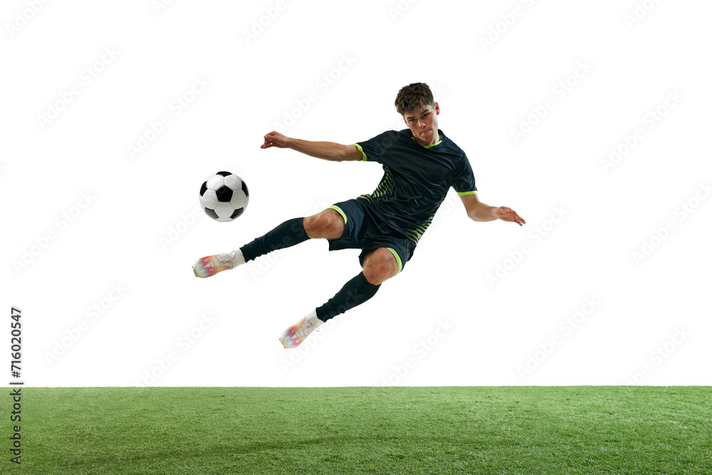 Obraz premium Young man, soccer player in black uniform, hitting ball in a jump, playing isolated over white background with grass flooring. Concept of sport, game, competition, championship, active lifestyle