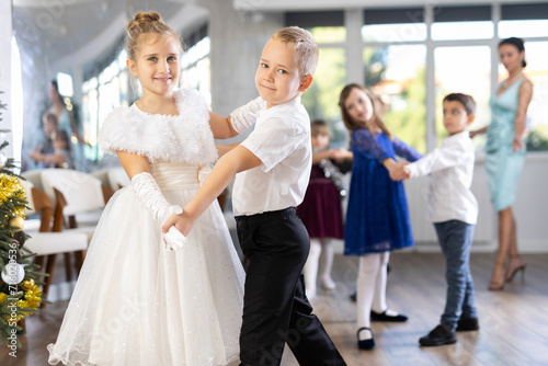 Smiling junior schoolchildren in party dresses learning dancing waltz with pedagogue in social hall