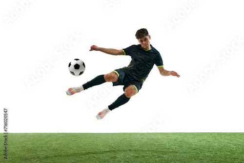 Young man, soccer player in black uniform, hitting ball in a jump, playing isolated over white background with grass flooring. Concept of sport, game, competition, championship, active lifestyle