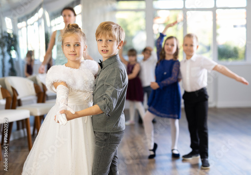 Happy interested preteen pupils preparing for festive school event, rehearsing ballroom dance steps in spacious classroom while female teacher observing from background..
