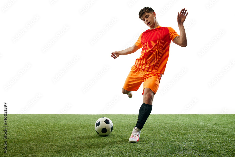 Young man, football player in orange uniform playing, training, hitting ball isolated over white background with grass flooring. Concept of sport, game, competition, championship, active lifestyle