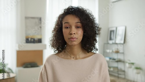 Portrait confused woman of color asking question, frowning brows uncertainty. Hesitant multi ethnic elegant lady in modern interior home. Unconfident young biracial woman talking to camera living room photo