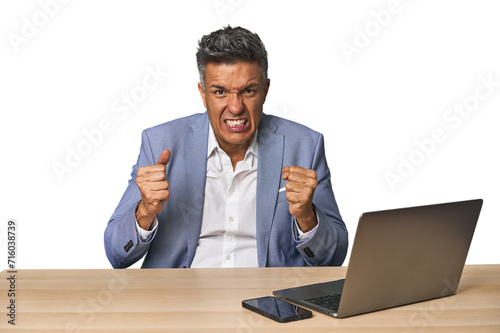 Elegant businessman at desk with laptop upset screaming with tense hands.