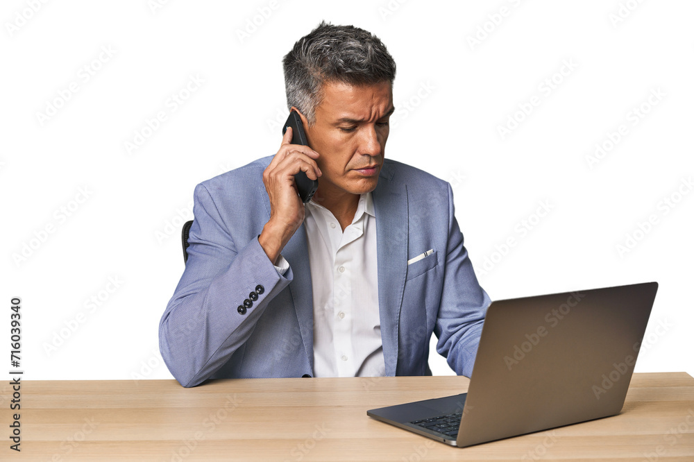Serious middle-aged Hispanic man on phone while working on laptop Serious middle-aged Hispanic man on phone while working on laptop