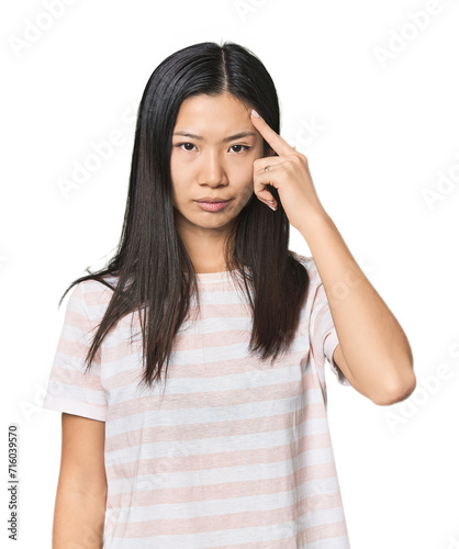 Young Chinese woman in studio setting pointing temple with finger, thinking, focused on a task.