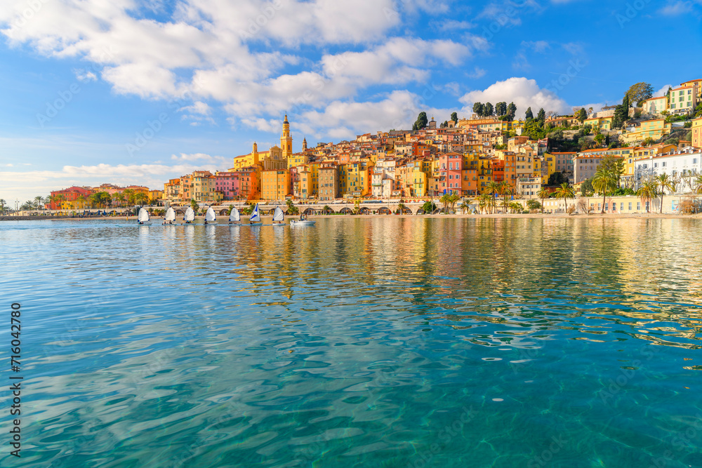 A small group of sailboats pass in front the old town and Les Sablettes Beach and promenade along the Cote d'Azur French Riviera at Menton, France.