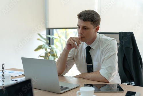 Focused man in a tie thinks while working on his laptop in a sunny office with a coffee cup and papers.