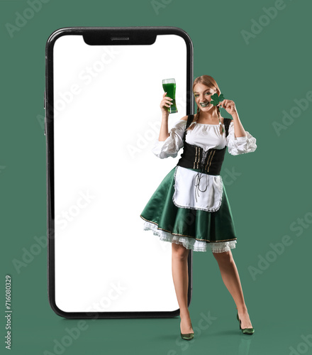 Beautiful Irish waitress with glass of beer and clover on green background. St. Patrick's Day celebration