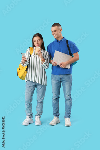 Happy students with backpacks, cup of coffee and laptop using mobile phone on blue background