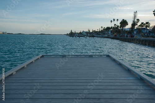 View looking south on edge of pier toward sea wall east of Pass A Grille Way into Pass-a-Grille Channel near sunset with calm waves, blue and white sky.