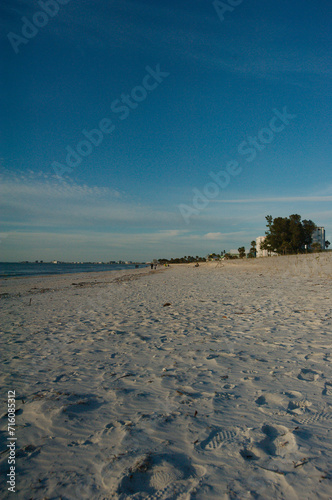 Vertical low angle view over sand of Pass-s-grille beach in St. Pete Beach Florida looking north. Near sunset with shadows in sand and white clouds. Green trees and buildings on right side.