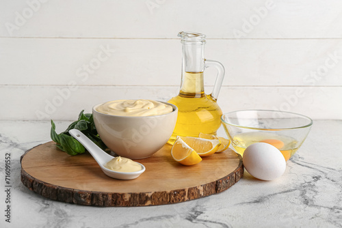 Bowl and spoon of fresh mayonnaise with ingredients on table near white wooden wall
