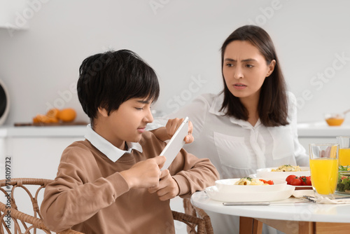 Angry mother taking tablet computer away from her teenage son at table during dinner in kitchen. Family problem concept