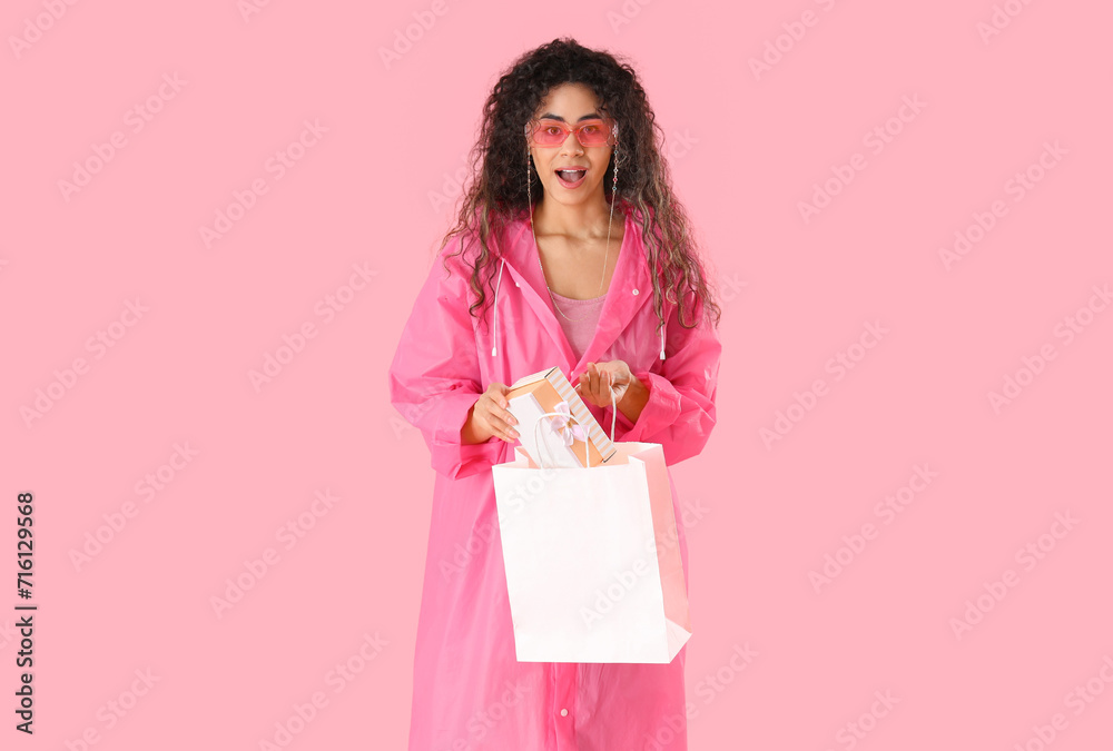 Surprised African-American woman with gift box and shopping bag on pink background. International Women's Day celebration Surprised African-American woman with gift box and shopping bag on pink background. International Women's Day celebration