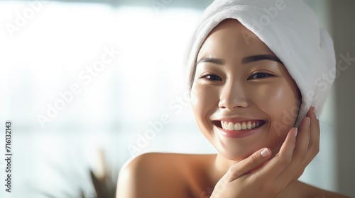 A Closeup head shot of a young beautiful Asian woman applying facial moisturizer after bathing. Smiling beautiful woman wrapped in towel to smooth her skin Daily routine in the morning. photo