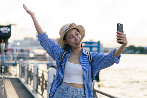 Traveler asian young woman using smart phone camera to take a selfie photo, selfie at riverside of Chaopraya River in Bangkok. Backpacker travel on holiday trip or vacation concept