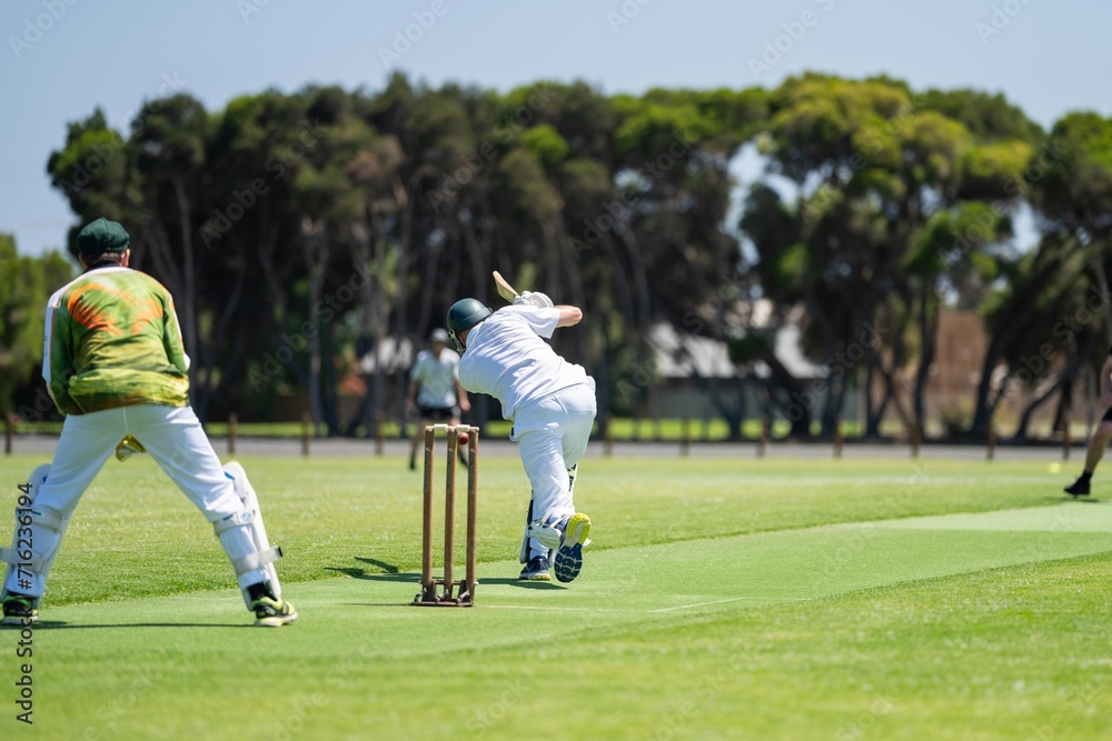 a local cricket match being played on a green cricket oval in summer in australia. australian cricketer batting and bowling in a game