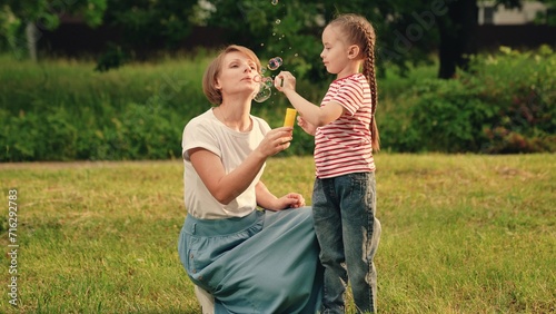 Little girl asks mother to blow soap bubbles in urban park. Happy mother enjoys blowing bubbles with daughter resting in grassy meadow. Cute child with braids spends time with mommy on sunny day. Kid