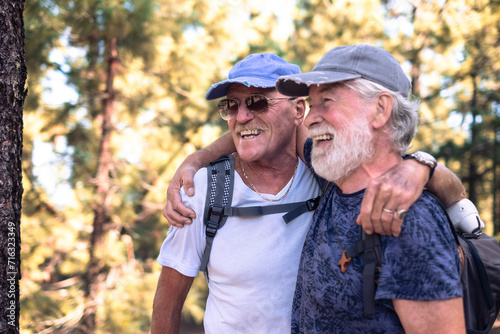 Cheerful couple of old active men with hat and backpack embrace have fun in mountain hike in the woods appreciating adventure and freedom, retired seniors and healthy lifestyle concept