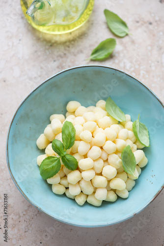 Turquoise bowl with raw italian chicche di patate and fresh green basil, vertical shot on a beige granite surface, elevated view