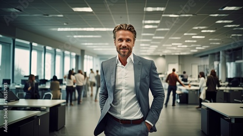 Smiling man in business attire standing confidently in a bustling modern office with team members in the background.