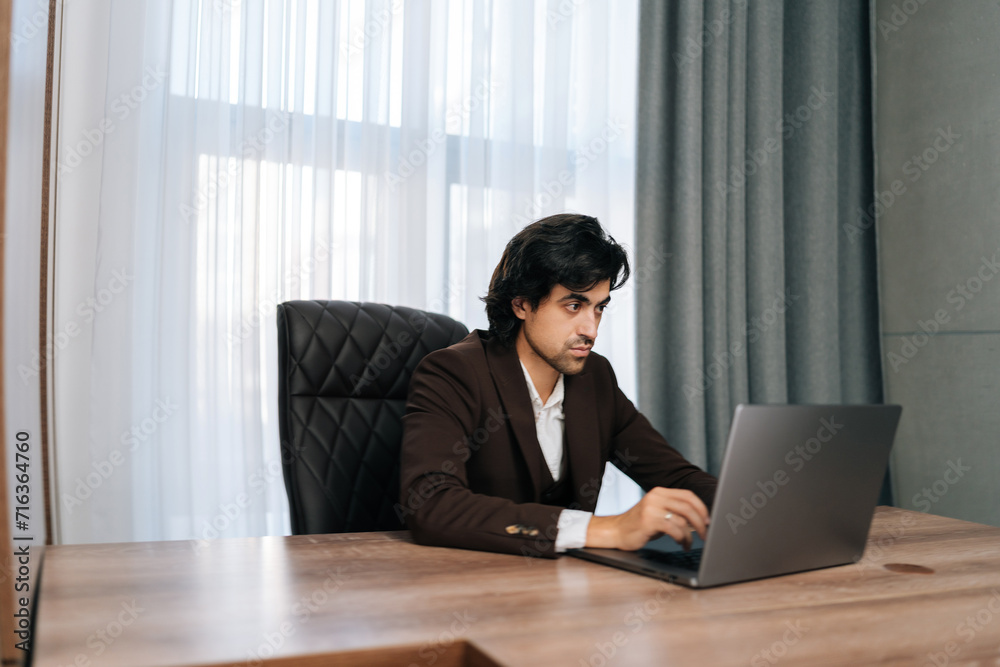 Side view of bearded businessman sitting at workplace by window, working on laptop, looking at screen, typing messages. Executive CEO dressed in suit concentrate working with notebook at workplace. Side view of bearded businessman sitting at workplace by window, working on laptop, looking at screen, typing messages. Executive CEO dressed in suit concentrate working with notebook at workplace.