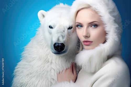 A blonde woman in a fur coat with a hood hugs a polar bear. Portrait on a blue background
