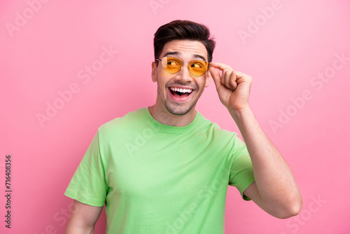 Photo of young macho smiling guy trying new sunglasses oakley wearing green t shirt looks empty space isolated on pink color background photo
