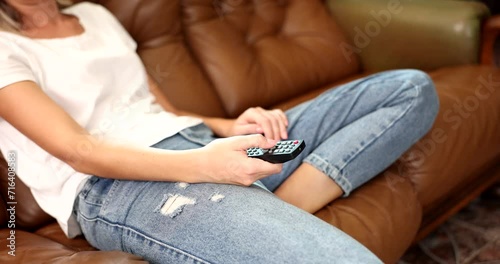 Bored woman sitting on couch holding TV remote control and switching channels. Person looking uninterested selects content photo