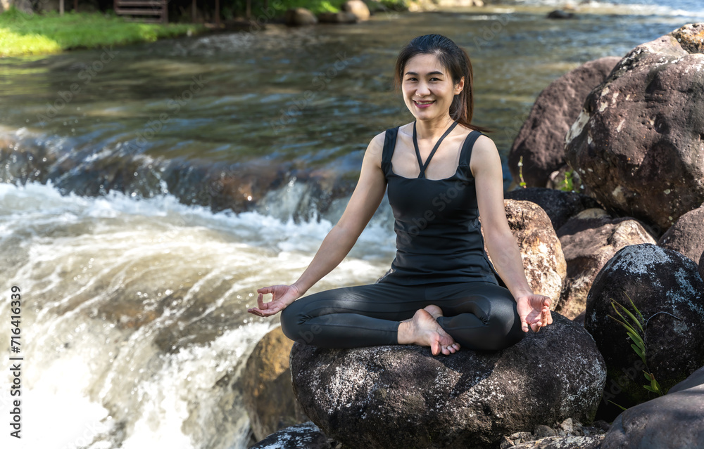 Happy Asian woman sitting on rock near river doing lotus pose yoga meditating Happy Asian woman sitting on rock near river doing lotus pose yoga meditating