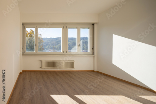 Empty room with white walls and a large  bright window that lets in direct sunlight. Outside the window are trees and hills