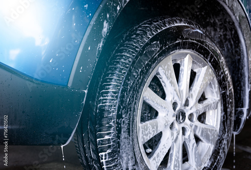 Blue car wash with white soap foam and professional auto care service. Car cleaning service concept. Vehicle cleaning service. Foam wash car detailing. Tire of car wheel is covered with white foam.