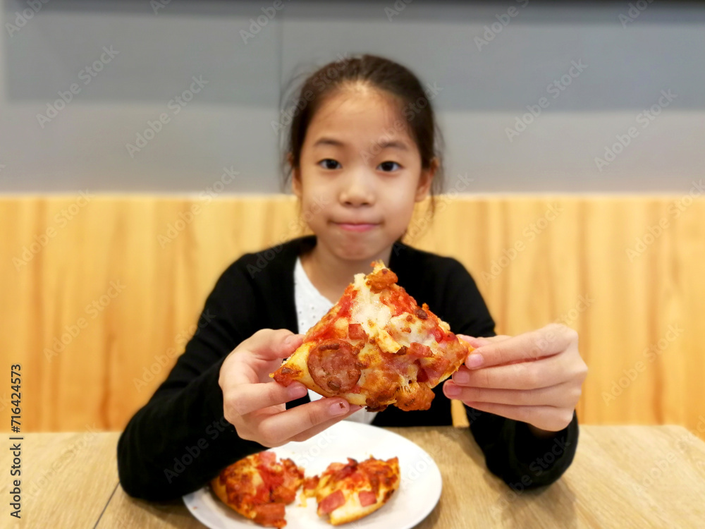 Happy Asian child enjoy eating pizza on table. Portrait of a young girl with an appetizing piece of pizza. Thai girl eating pizza at cafe, concept for unhealthy junk food. Focus on pizza. Happy Asian child enjoy eating pizza on table. Portrait of a young girl with an appetizing piece of pizza. Thai girl eating pizza at cafe, concept for unhealthy junk food. Focus on pizza.