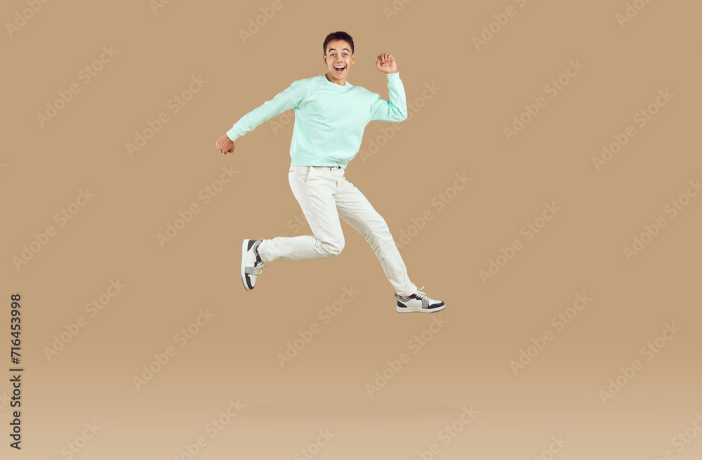 Happy cheerful excited young man or teenage boy in casual mint sweatshirt and white pants running and jumping high in air isolated on light brown color background