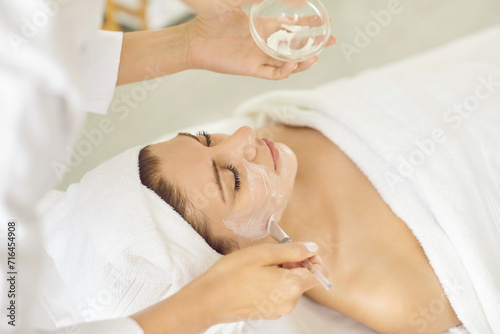 Cosmetologist applying moisturizing mask with brush to a young relaxed smiling young woman in spa salon. Close up portrait of a girl client receiving beauty procedure from beautician in beauty clinic photo