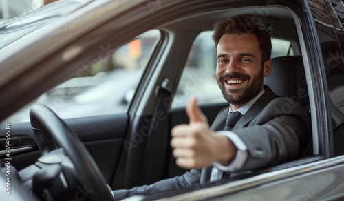 Side view of businessman in suit driving expensice car, smiling happily and showing thumb up, closeup. Young man entrepreneur going home by car after successful business meeting. © radekcho