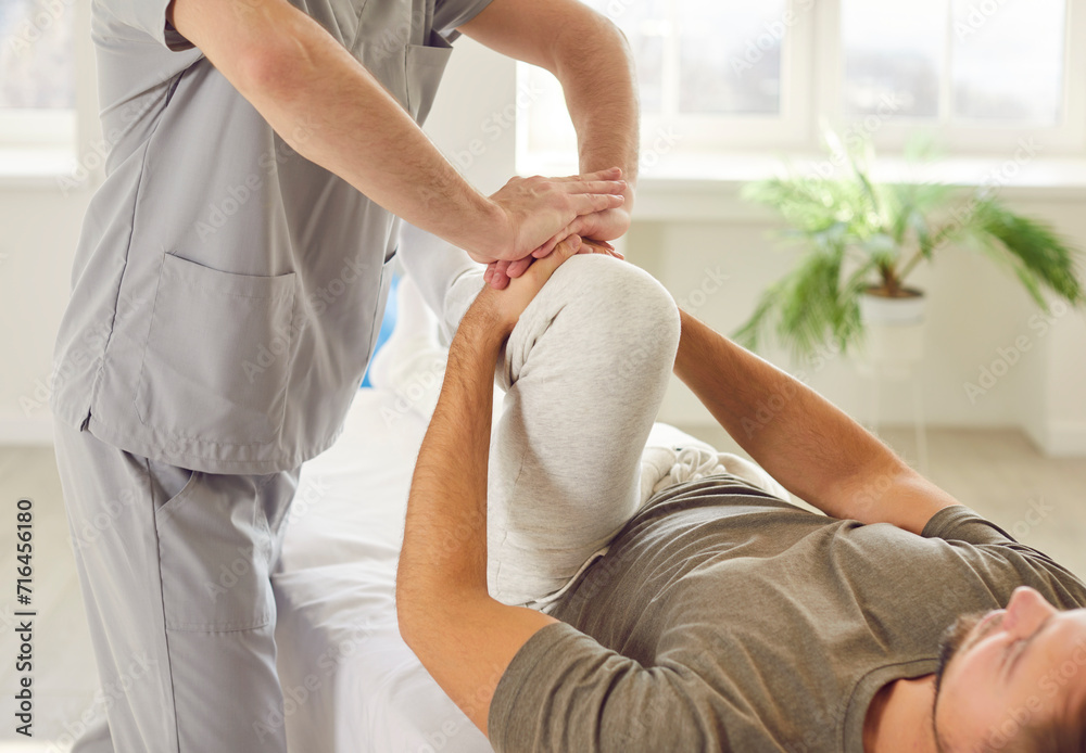 Male physical therapist doing healing treatment on mans knee in rehabilitation clinic. Professional physiotherapist or osteopath working in office. Physiotherapy and osteopathic medicine concept. - obrazy, fototapety, plakaty 