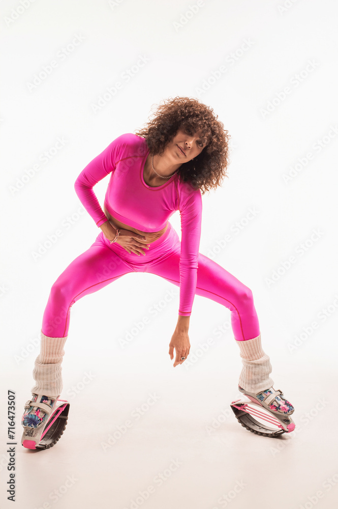 Sporty woman with curly hair in pink tracksuit wearing kangoo jumpers on studio background
