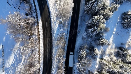 Aerial shot of a truck driving on a winding asphalt road through a snowy forest ion a sunny winter day.