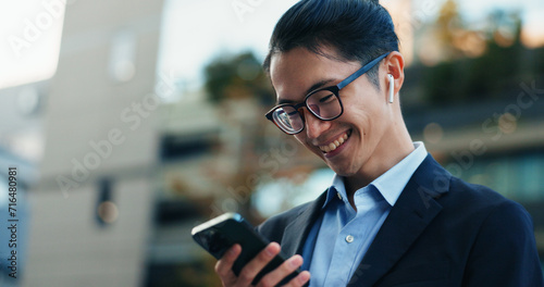 Phone, smile and Japanese businessman in the city reading company email on technology. Happy, career and professional young male person scroll or browse on cellphone with earbuds in urban town. photo