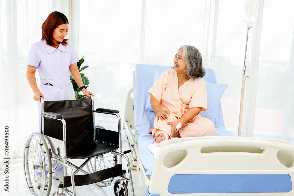 Obraz premium Lovely, kind-hearted female nurse brings wheelchair to move and care for an elderly female patient with ankle pain from sprained foot uses bandage to take her to be examined by medical professional.