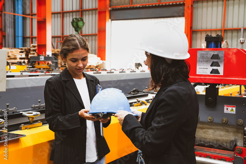 Female factory manager gladly handed over safety helmet while taking female business woman and business partner on a tour of the production work area in the metal sheet industry to ensure safety.