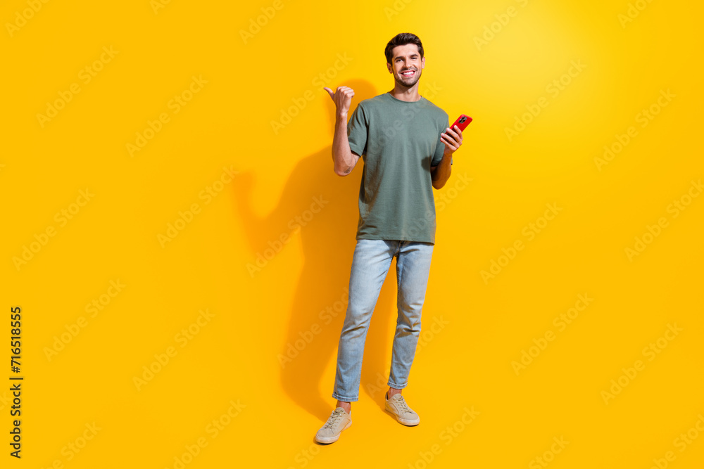 Full length photo of positive funny man dressed khaki t-shirt texting modern gadget thumb empty space isolated yellow color background Full length photo of positive funny man dressed khaki t-shirt texting modern gadget thumb empty space isolated yellow color background