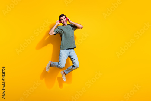 Full length photo of cheerful dreamy man dressed khaki t-shirt enjoying music earphones jumping empty space isolated yellow color background