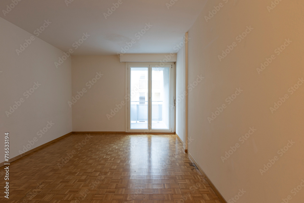 Front view of an empty room with white walls and parquet. There is a window overlooking the balcony
