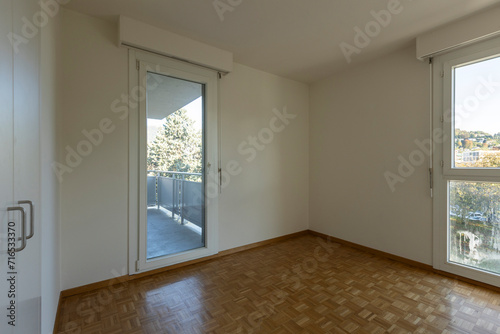 Empty room with white walls and parquet flooring. There are two windows, one leading to the balcony, the other to the sunlit countryside