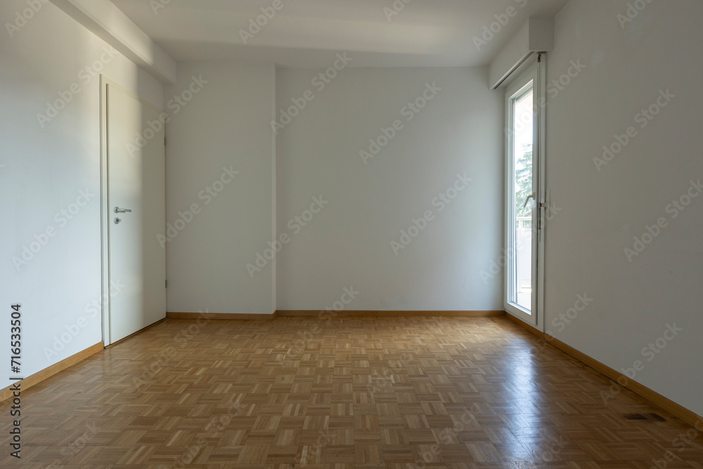Front view of an empty room with parquet flooring and white walls. On the left is a window letting in a lot of natural light, on the right is a closed door. Front view of an empty room with parquet flooring and white walls. On the left is a window letting in a lot of natural light, on the right is a closed door.