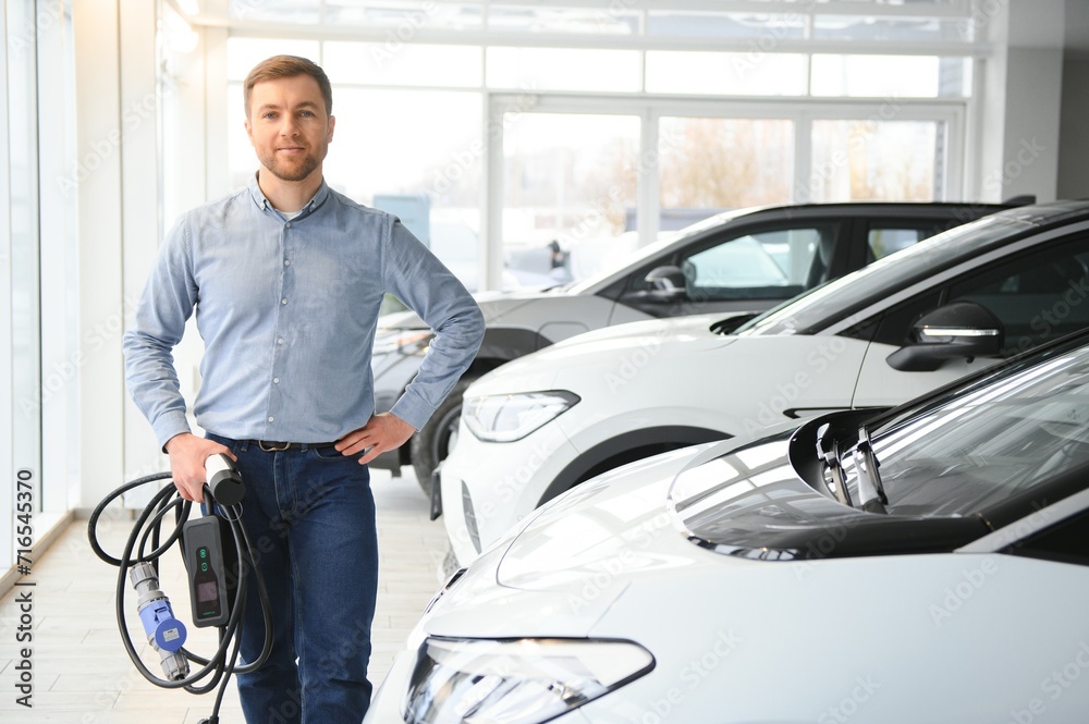 Young man buying first electric car in the showroom. Eco car sale concept